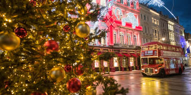 a christmas tree in the foreground, and the bus in the background lit up with fairy lights sits on a Christmas light decorated area of London