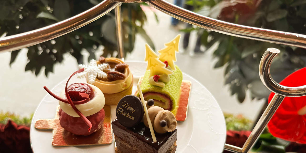 Four Christmas themed pastries, one green with white chocolate Christmas trees, one red, one tart with a snow flake and a layered chocolate pastry with 'merry Christmas' on it